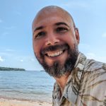 A young man with a beard smiling into the camera with a beach and the ocean in the background.