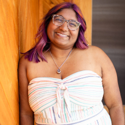 Woman leaning on a wooden wall with purple coloured hair and a strippy top. She is wearing glasses and smiles into the camer.