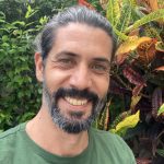 A man with his hair tied back in a green tshirt is smiling into the camera.